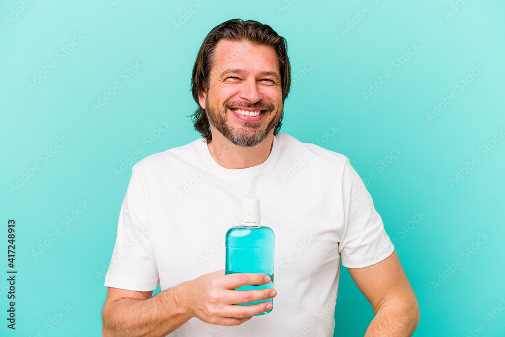 Middle age dutch man sitting holding a mouthwash isolated on blue background happy, smiling and cheerful.