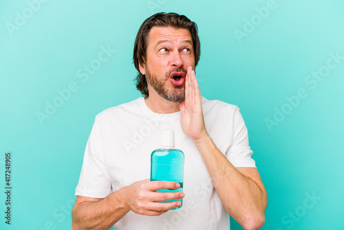 Middle age dutch man sitting holding a mouthwash isolated on blue background is saying a secret hot braking news and looking aside