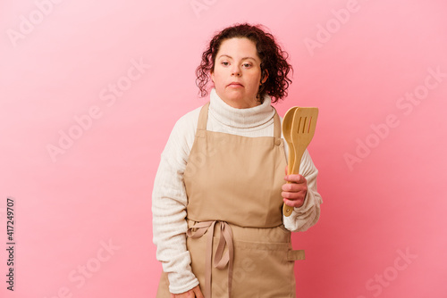 Woman with Down syndrome cooking at home isolated on pink background shrugs shoulders and open eyes confused.