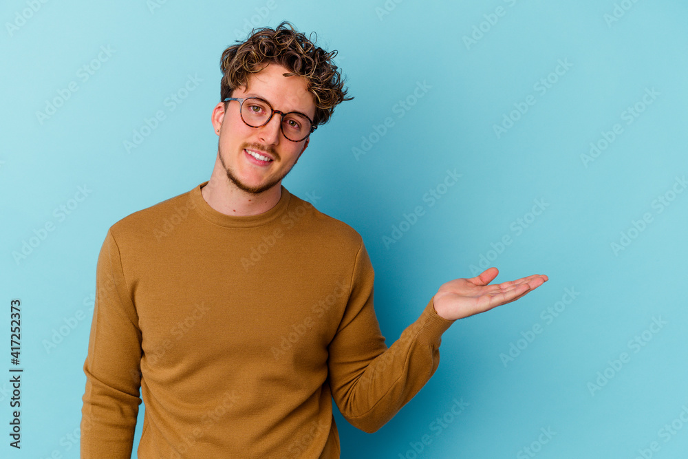 Young caucasian man wearing eyeglasses isolated on blue background showing a copy space on a palm and holding another hand on waist. Young caucasian man wearing eyeglasses isolated on blue background showing a copy space on a palm and holding another hand on waist.