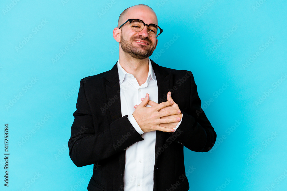 Young business caucasian bald man isolated on blue background has friendly expression, pressing palm to chest. Love concept. Young business caucasian bald man isolated on blue background has friendly expression, pressing palm to chest. Love concept.