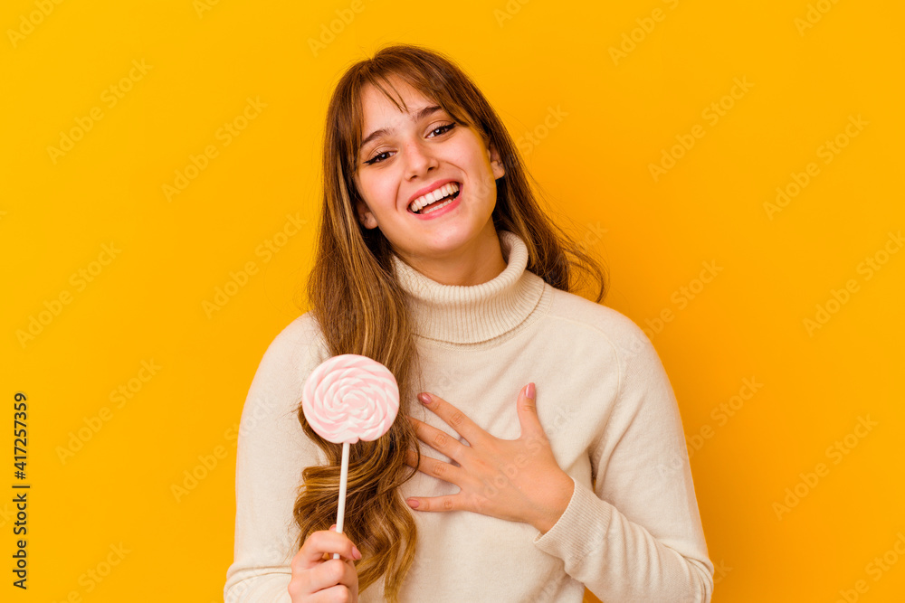 Young caucasian woman holding a lollipop isolated on yellow background laughs out loudly keeping hand on chest. Young caucasian woman holding a lollipop isolated on yellow background laughs out loudly keeping hand on chest.