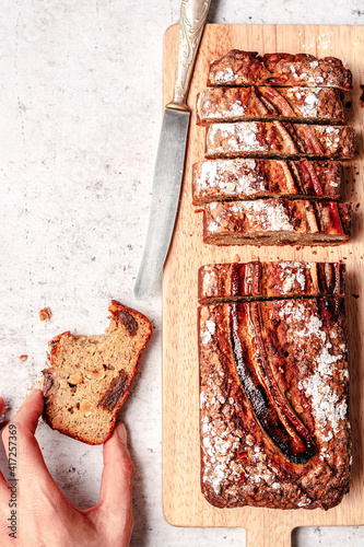 Whole grain banana bread with wallnuts and dried dates, sliced and served on a wooden cutting board with a hand grabbing one piece and a knife. photo