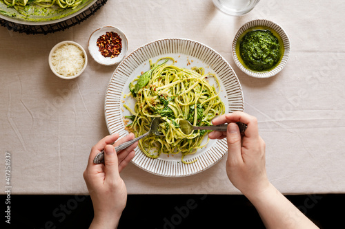 Hands twirling a plate of creamy pesto pasta next to a glass of water & small bowls with pasta toppings.