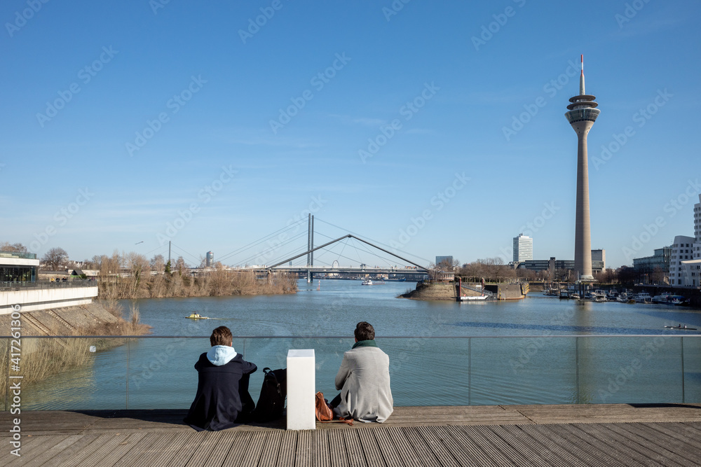 Sunny view, men sit on Brücke Im Medienhafen, pedestrian bridge at Media Hafen and background of harbour, Rhein tower, Neuer Zollhof and promenade on waterfront of Rhein River in Düsseldorf, Germany.
