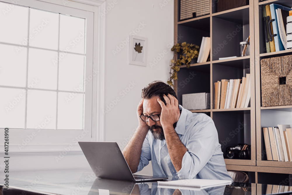 One alone unhappy and sad man at home in the office using laptop and looking at it with hands in the head. Stressed male in trouble resolving a problem One alone unhappy and sad man at home in the office using laptop and looking at it with hands in the head. Stressed male in trouble resolving a problem