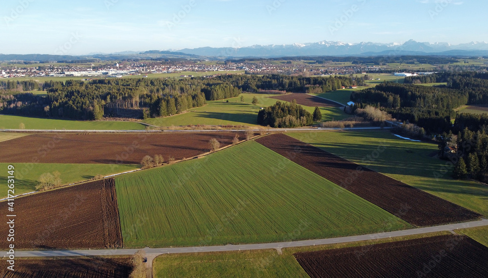 Luftaufnahme mit einer Drohne von verschiedenfarbigen Feldern, einer Straße und den Alpen im Frühling in Bayern