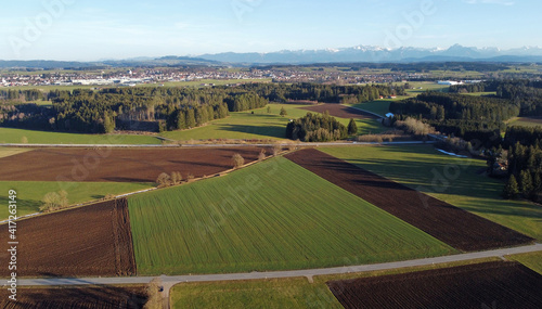 Luftaufnahme mit einer Drohne von verschiedenfarbigen Feldern, einer Straße und den Alpen im Frühling in Bayern