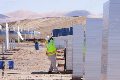 Male workers on construction site at solar farm pulling solar panel off tall stack of panels to install on metal support structure