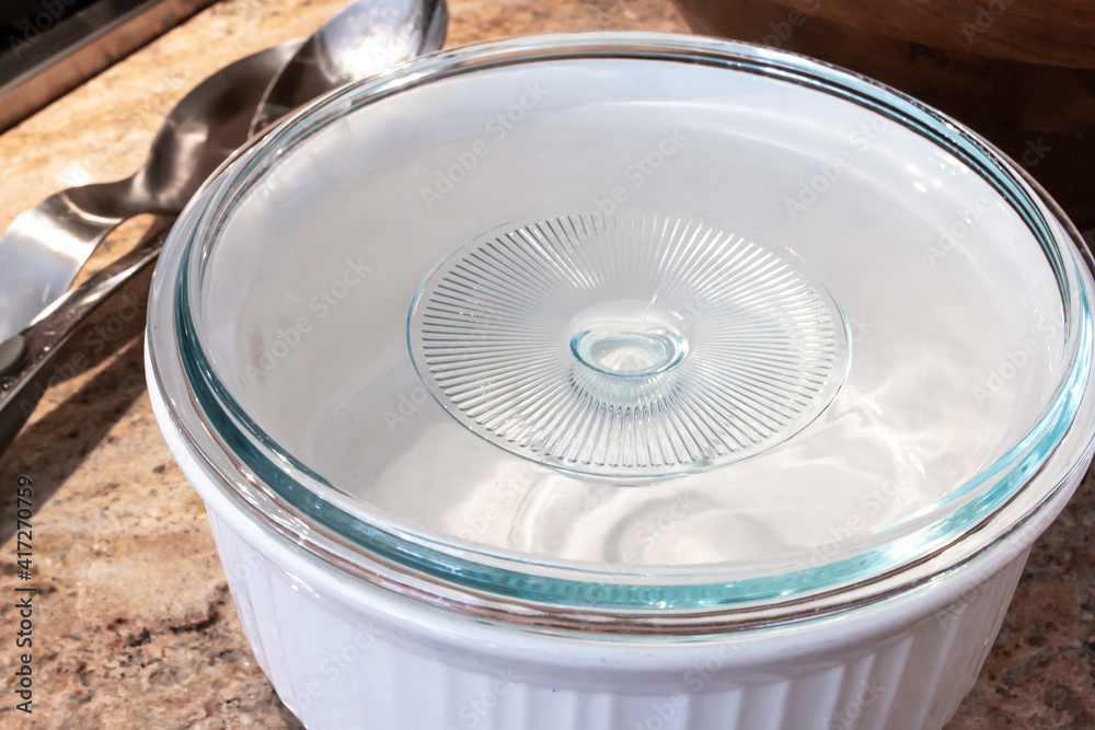 Empty white ceramic cooking pot with a clear glass lid sitting on top upside down. Shot on a marble countertop with two silver spoons next to the pot, 2021, Ontario Canada. Empty white ceramic cooking pot with a clear glass lid sitting on top upside down. Shot on a marble countertop with two silver spoons next to the pot, 2021, Ontario Canada.