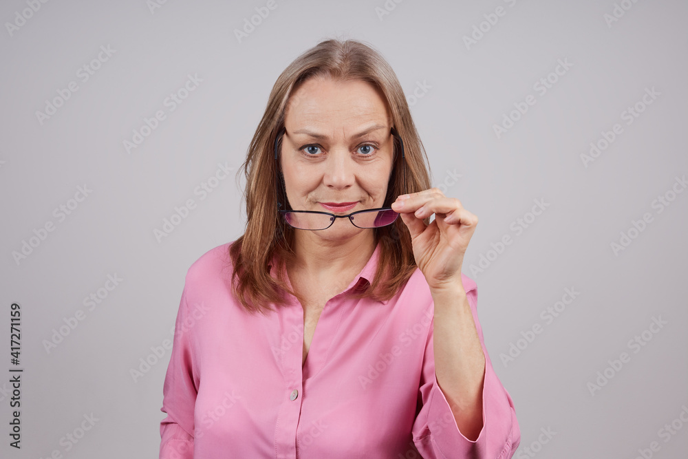 Nice mature businesswoman in pink blouse takes off glasses, looks over the top. Studio photo over gray background with copy space. Nice mature businesswoman in pink blouse takes off glasses, looks over the top. Studio photo over gray background with copy space.