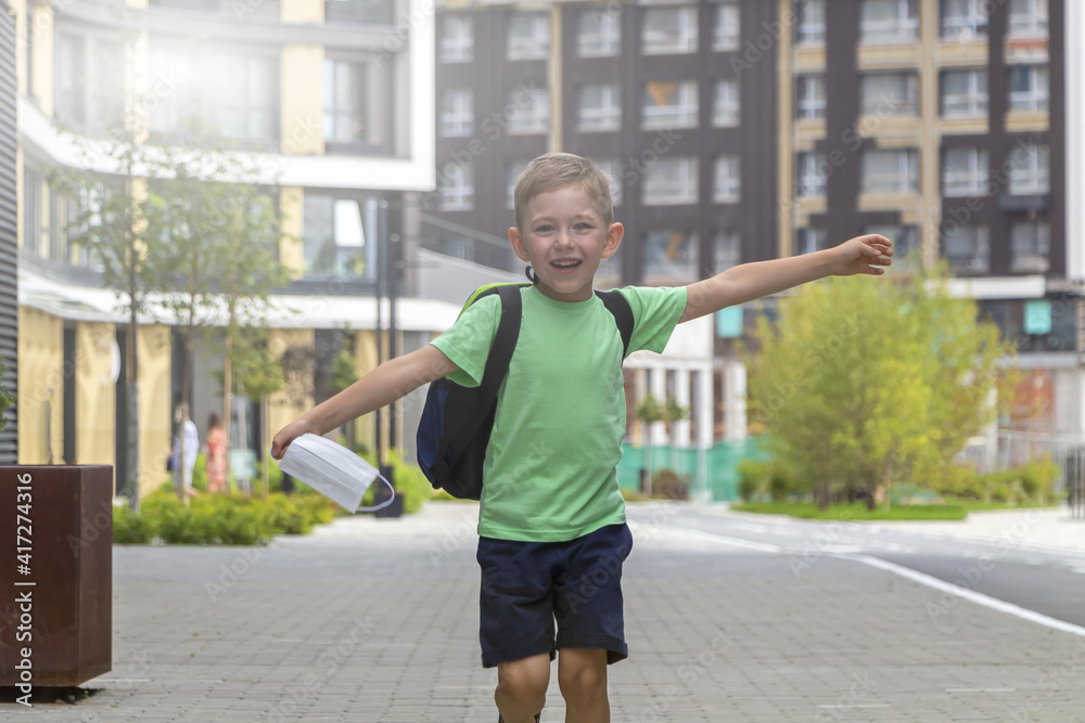 Happy smiling schoolboy with backpack and medical mask in his hand runs after end of lessons. New normal, back to school consept