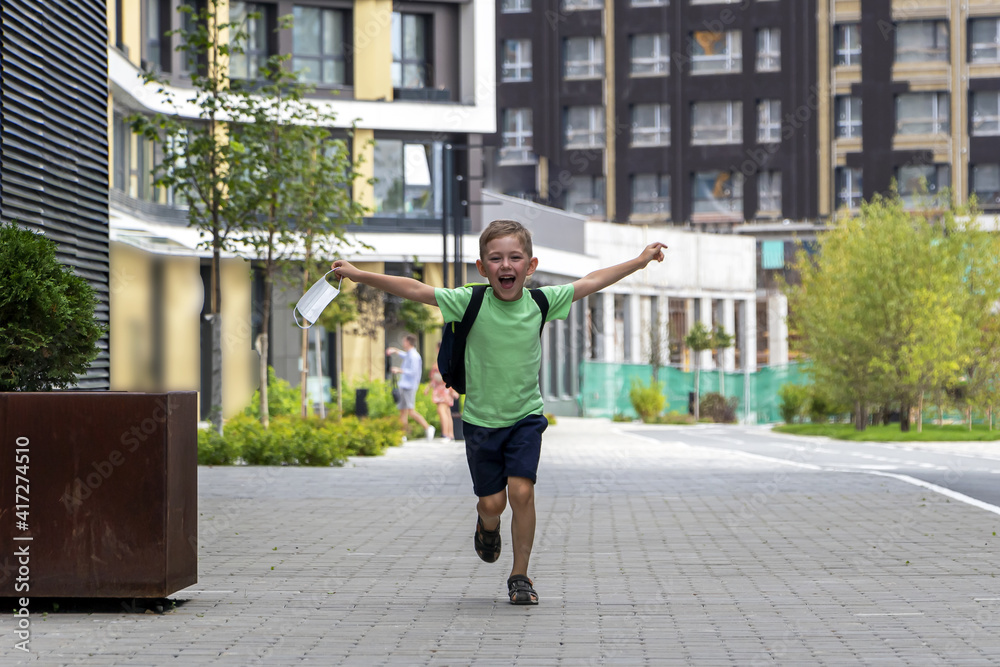 Happy smiling schoolboy with backpack and medical mask in his hand runs after end of lessons. New normal, back to school consept