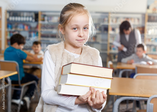 Portrait of happy cute preteen girl standing in classroom with books in hands ..