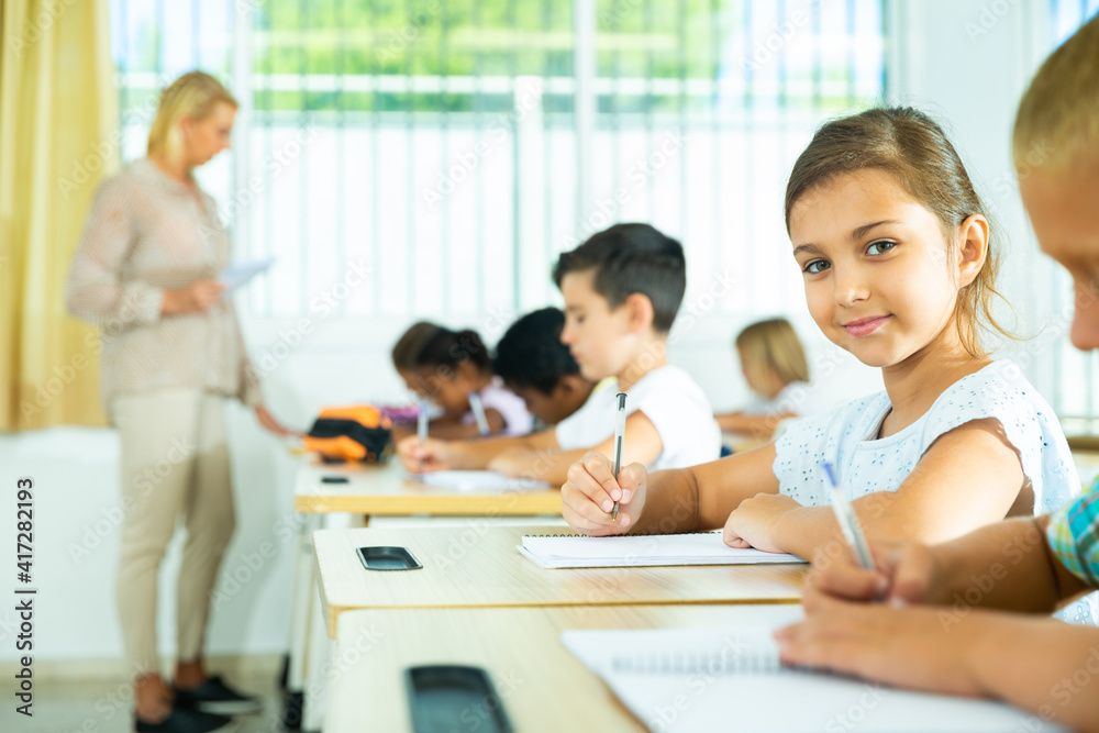 Focused girl sitting at desk writing test in classroom full of pupils during lesson