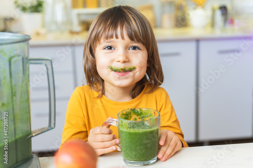 Happy Kid with glass cup of green smoothies in hands. Cute boy crazy drinks healthy dietary nutritious cocktail at home in the kitchen. Healthy lifestyle, raw food