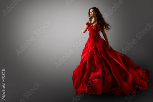 Fashion Woman in Red Dress. Beauty Model dancing in Long Evening Gown fluttering on Wind Black Hair flying in Air over Gray Background