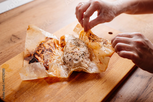 Baked chicken breast wrapped in parchment paper. A woman cuts a chicken breast baked with spices and herbs. The baked chicken fillet is cut into pieces on the table.
