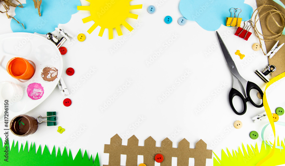 Fototapeta premium The background is a view from above on a white table with children's creativity. On the table of colored paper the sun, clouds, grass, fence, palette, scissors, colored buttons.