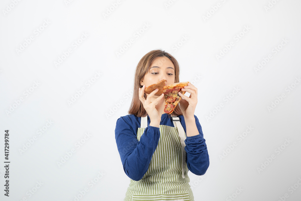 Portrait of young woman in apron eating slice of pizza on white background Portrait of young woman in apron eating slice of pizza on white background