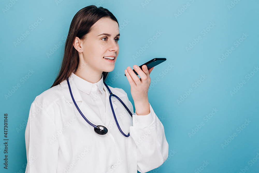 Smiling female doctor wearing white medical uniform and stethoscope holding phone speak activate virtual digital voice assistant on smartphone, consult patient online, isolated on blue background Smiling female doctor wearing white medical uniform and stethoscope holding phone speak activate virtual digital voice assistant on smartphone, consult patient online, isolated on blue background