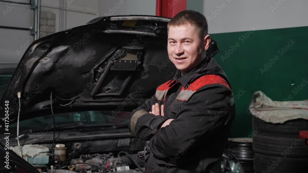 Cheerful technician with crossed arms in grey uniform stands near renovated automobile with open bonnet in workshop of car service station Cheerful technician with crossed arms in grey uniform stands near renovated automobile with open bonnet in workshop of car service station