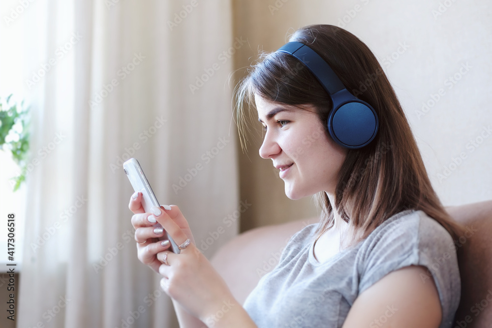 Happy girl sitting at home on the couch and listening to music from the application on the phone with headphones - Young millennial woman resting and listening to an online training course or podcast Happy girl sitting at home on the couch and listening to music from the application on the phone with headphones - Young millennial woman resting and listening to an online training course or podcast