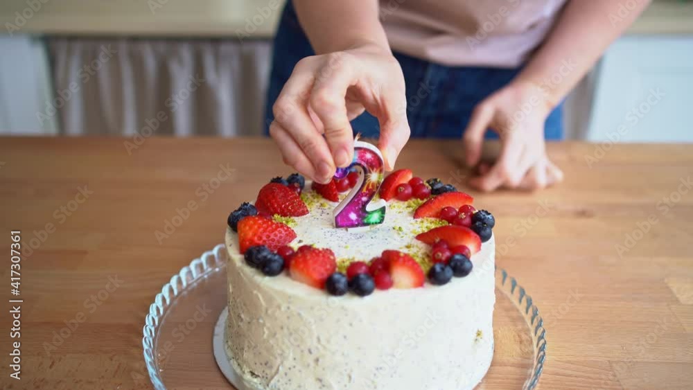 A woman in the kitchen puts a candle in the shape of the number 2 on the cake A woman in the kitchen puts a candle in the shape of the number 2 on the cake