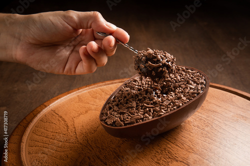 Wall Mural Hand sticking a metallic spoon in a stuffed chocolate easter egg with grated chocolate on the top on a wooden stand on a wooden table