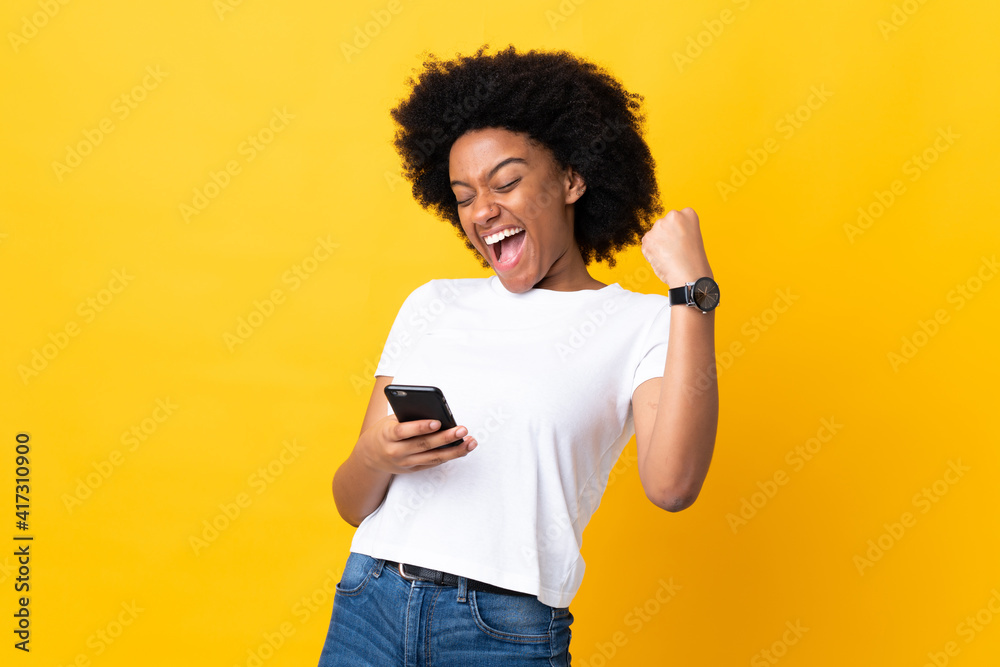 Young African American woman isolated on yellow background with phone in victory position