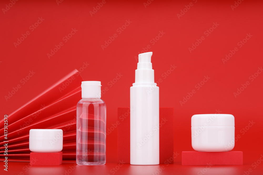 Set of white unbranded cosmetic bottles with lip balm, skin lotion, face cream and hydrating mask or scrub against red background decorated with red waver. Concept of luxury or asian style skin care Set of white unbranded cosmetic bottles with lip balm, skin lotion, face cream and hydrating mask or scrub against red background decorated with red waver. Concept of luxury or asian style skin care