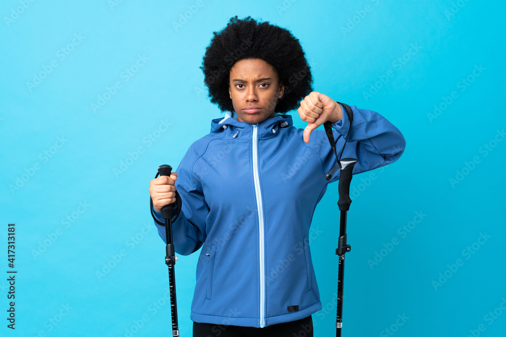 Young Africa American with backpack and trekking poles isolated on blue background showing thumb down with negative expression