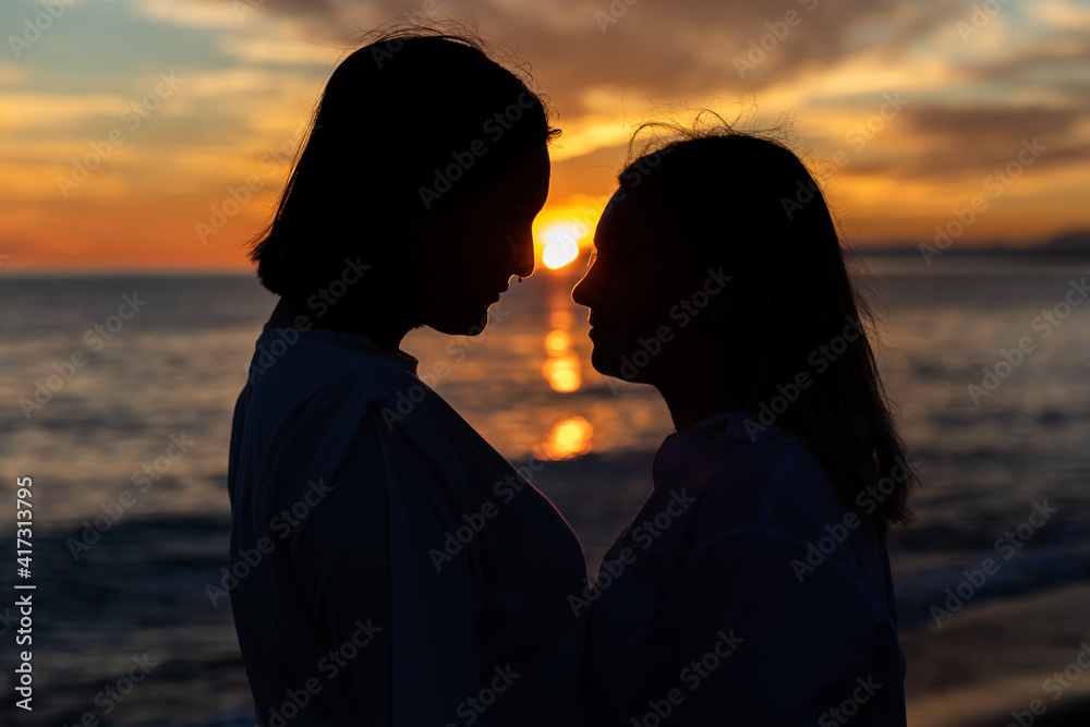 Joven pareja lesbiana mirándose bajo un atardecer a través de una silueta oscura con precioso mar y fondo de nubes con sol