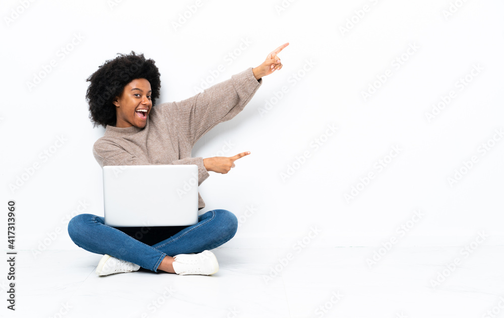 Young African American woman with a laptop sitting on the floor pointing finger to the side and presenting a product Young African American woman with a laptop sitting on the floor pointing finger to the side and presenting a product