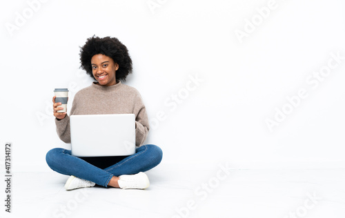 Young African American woman with a laptop sitting on the floor holding coffee to take away and a mobile © luismolinero