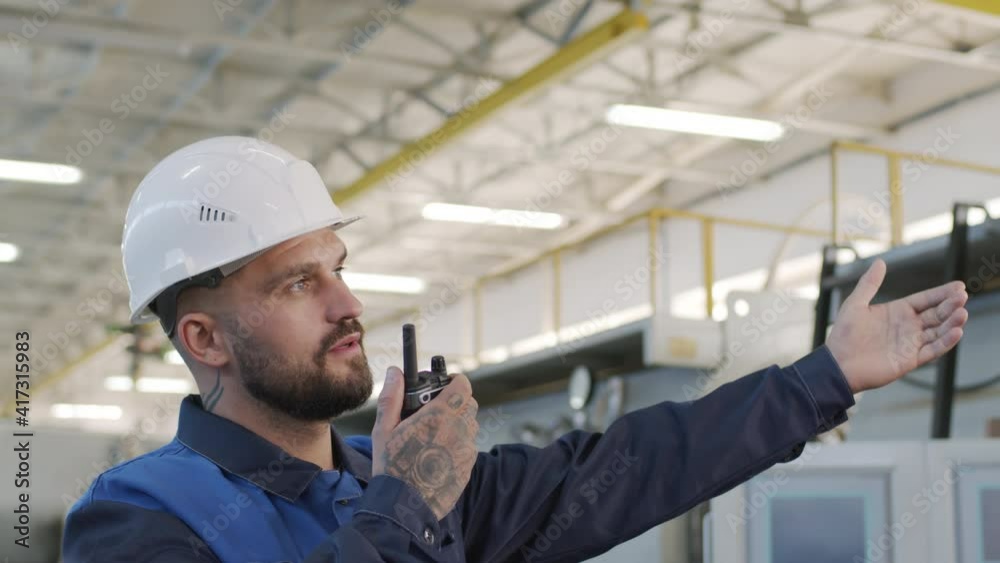 Slowmo of bearded male plant supervisor in hard hat talking on walkie-talkie and pointing at something Slowmo of bearded male plant supervisor in hard hat talking on walkie-talkie and pointing at something