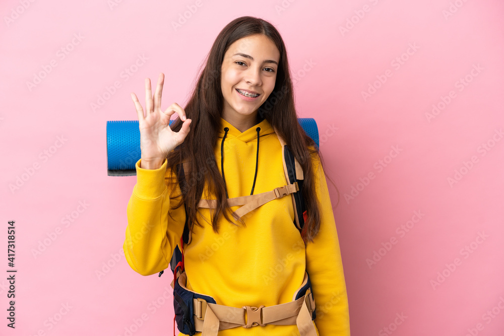 Young mountaineer girl with a big backpack isolated on pink background showing ok sign with fingers Young mountaineer girl with a big backpack isolated on pink background showing ok sign with fingers