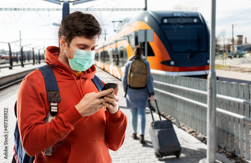 travel, tourism and pandemic concept - young man with smartphone and backpack wearing face protective medical mask traveling by train on railway station in city of tallinn, estonia on background