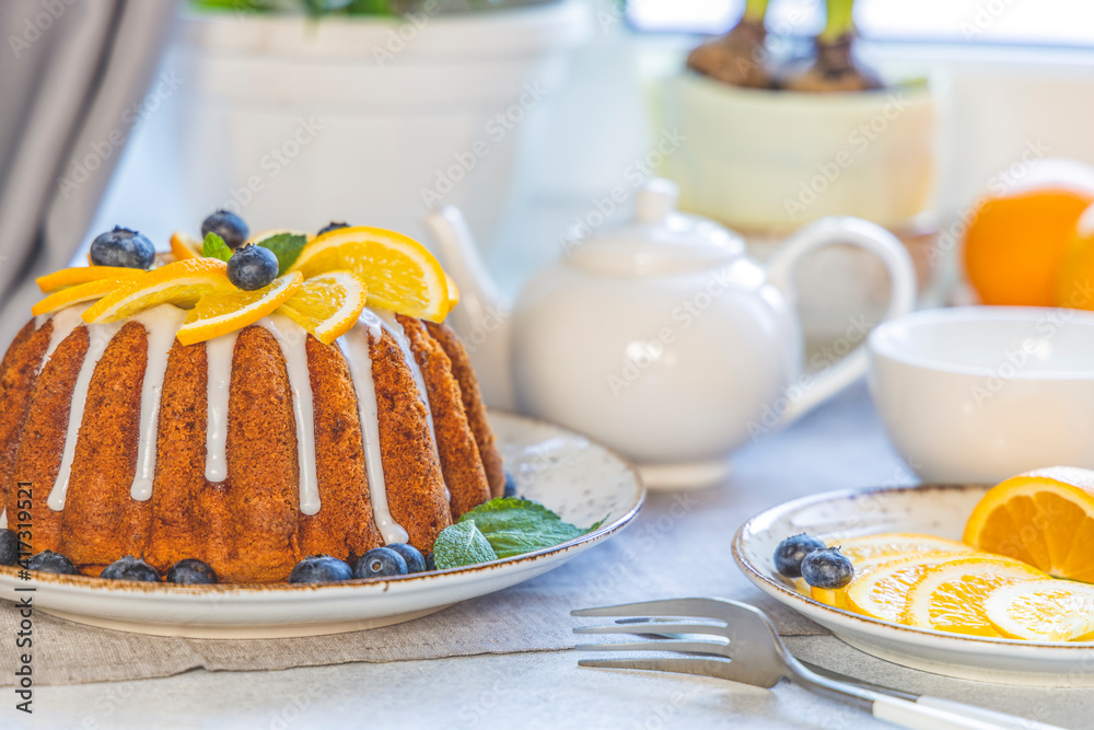 Orange bundt cake with blueberry surrounded fruits, plant and cutlery on light table near window. Family breakfast concept Orange bundt cake with blueberry surrounded fruits, plant and cutlery on light table near window. Family breakfast concept