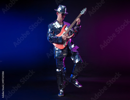 a male artist in a mirrored suit and hat with an electric guitar on a dark background
