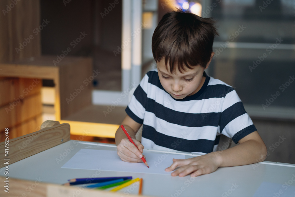adorable caucasian boy of elementary age drawing a rainbow with pencils sitting at the desk in his room at home. Image with selective focus adorable caucasian boy of elementary age drawing a rainbow with pencils sitting at the desk in his room at home. Image with selective focus