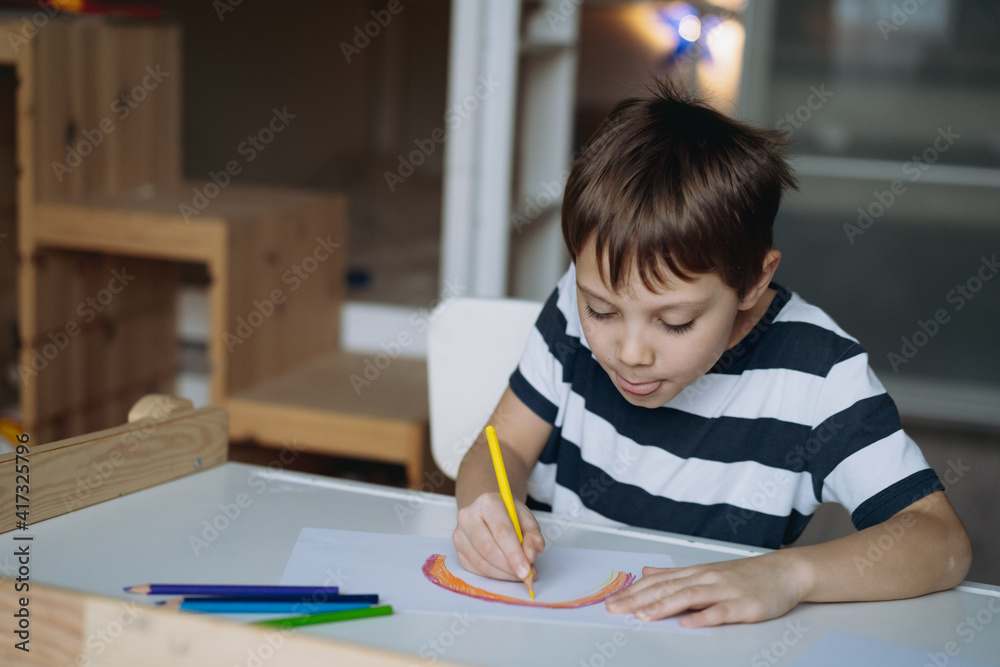adorable caucasian boy of elementary age drawing a rainbow with pencils sitting at the desk in his room at home. Image with selective focus