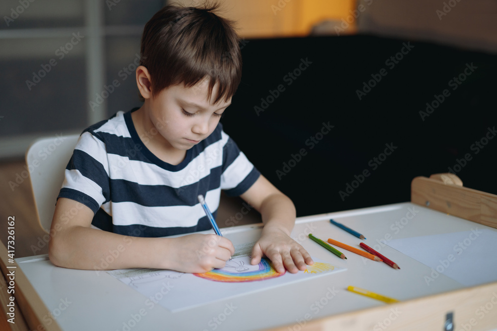 adorable caucasian boy of elementary age drawing with pencils sitting at the desk in his room at home. Image with selective focus adorable caucasian boy of elementary age drawing with pencils sitting at the desk in his room at home. Image with selective focus