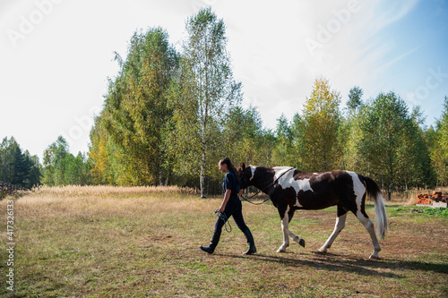 Along the forest lawn, a girl leads her horse by the bridle on a sunny autumn day.