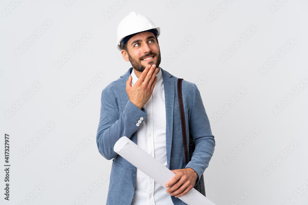 Young architect man with helmet and holding blueprints isolated on white background looking up while smiling