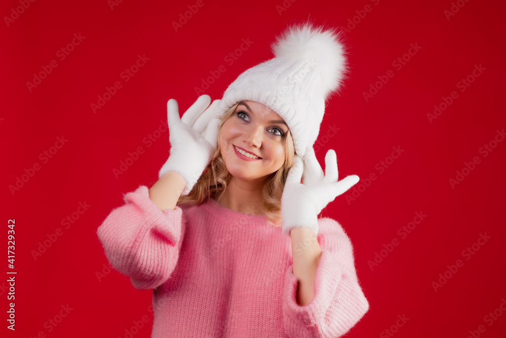 A cheerful blonde in a warm pink sweater and a white hat with gloves on a red background. Young woman in warm clothes, studio portrait. A cheerful blonde in a warm pink sweater and a white hat with gloves on a red background. Young woman in warm clothes, studio portrait.
