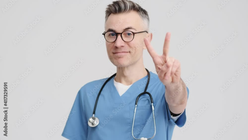 A close-up view of a happy handsome doctor man wearing uniform is showing peace gesture to the camera standing isolated over grey background in the studio A close-up view of a happy handsome doctor man wearing uniform is showing peace gesture to the camera standing isolated over grey background in the studio