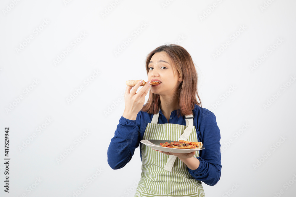 Portrait of female cook in apron eating slice of pizza on white background Portrait of female cook in apron eating slice of pizza on white background