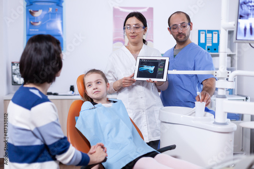 Dentist and patient commenting cavity treatment in tablet application after consultation. Stomatologist explaining teeth diagnosis to mother of child in health clinic holding x-ray.