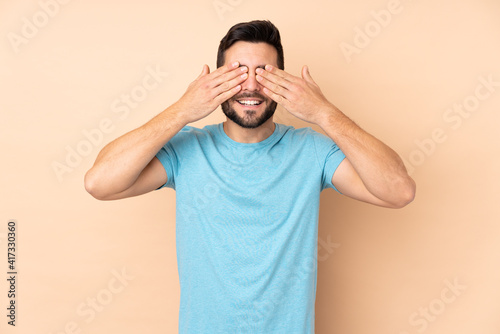 Caucasian handsome man isolated on beige background covering eyes by hands and smiling